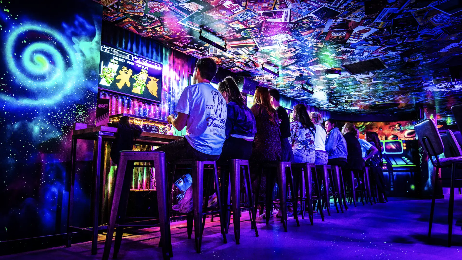 Row of arcade game machines being played by people at NQ64 Northern Quarter with colourful ceiling and wall decals