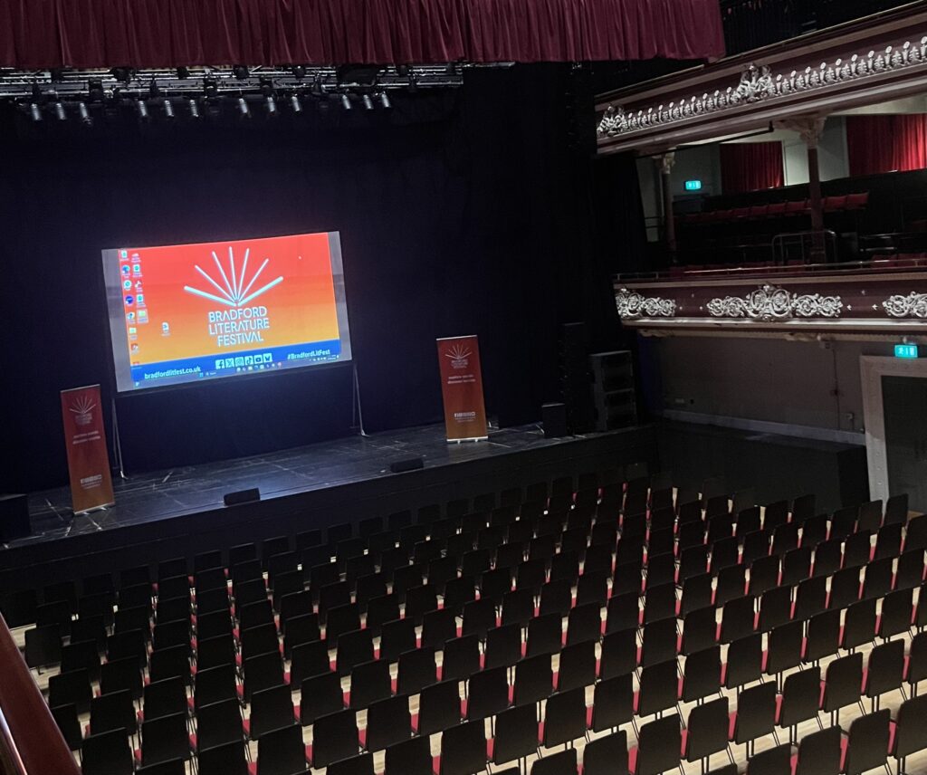 Theater interior. On stage is Bradford Lit Festival's logo projected onto the back.