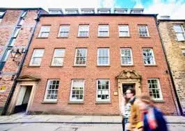 Front view of the Durham University History Department building, a red-brick Georgian-style structure with three floors of white-paned windows. Two people walk past in the foreground, slightly blurred in motion, under a bright sky with scattered clouds.