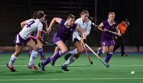 Durham University Women's Hockey. A match is being played at night. lPlayers in white and purple chase the ball on an astroturf pitch.