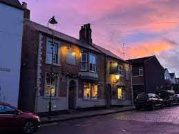 A double fronted Georgian pub, Ye Old Elm Tree, is lit from within. Behind the building, the sky is pink, orange, and blue from the sunset. 