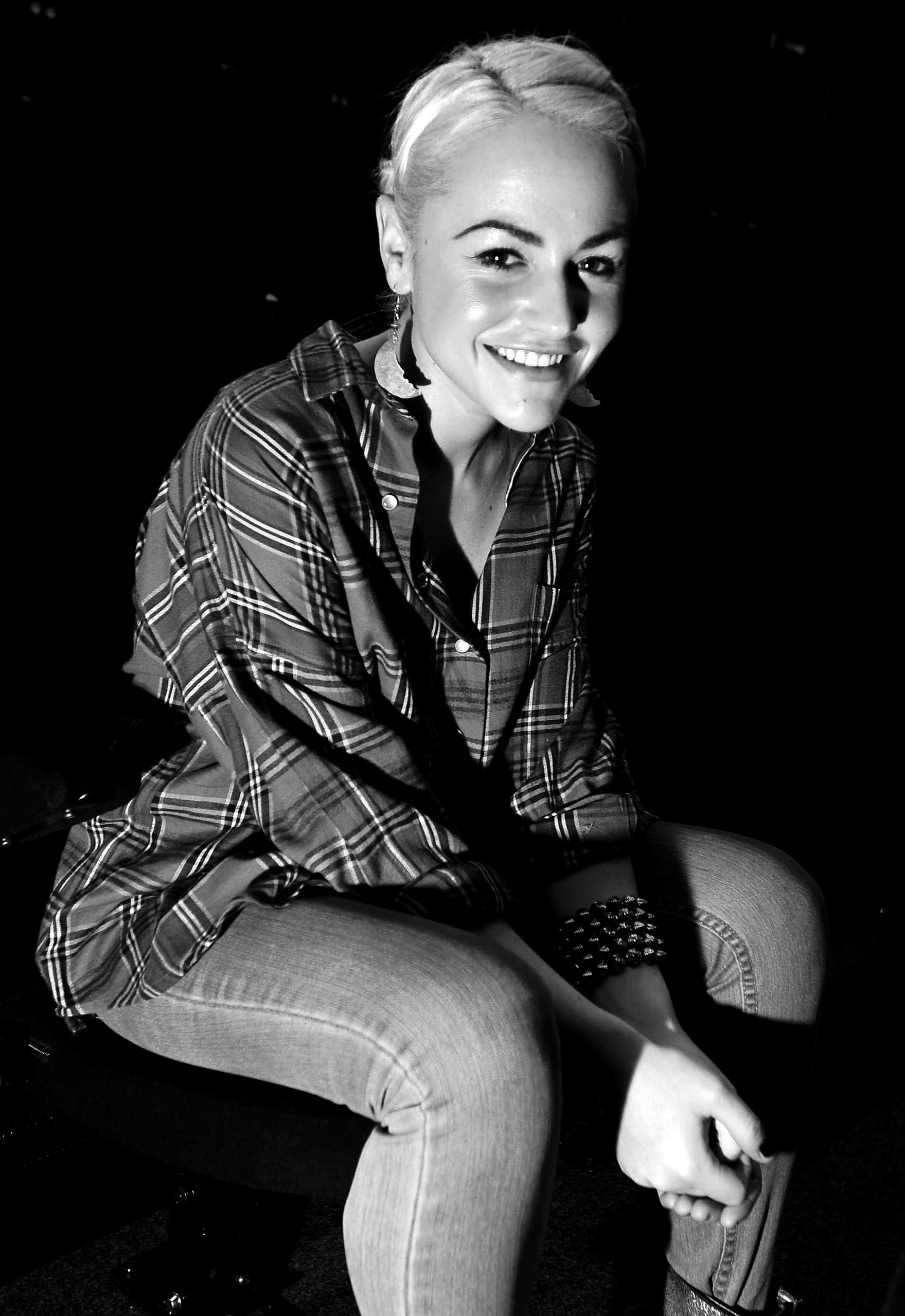 Black and white photo of actress Jaime Winstone smiling, seated, wearing a checked shirt